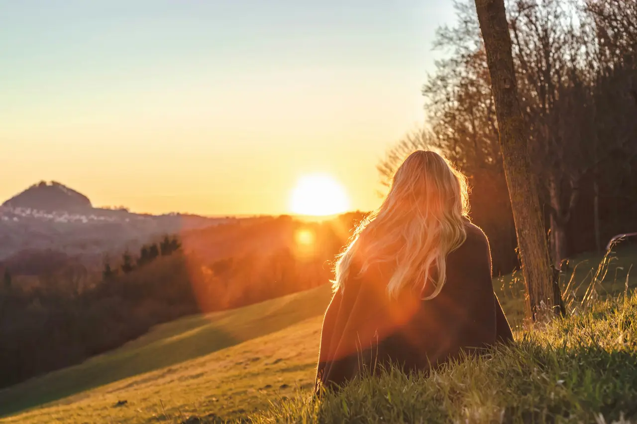 A woman sitting on a grassy hillside, facing the warm evening sun